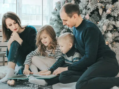 girl and boy reading book sitting between man and woman beside Christmas tree