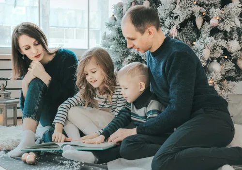 girl and boy reading book sitting between man and woman beside Christmas tree